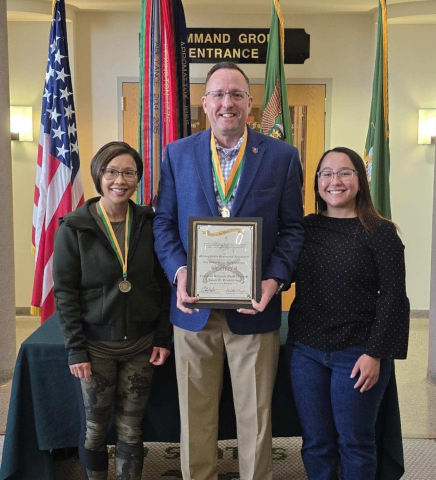image of James Breckinridge wearing his OoM in gold medal, holding an award certificate, beside him are his wife and daughter.